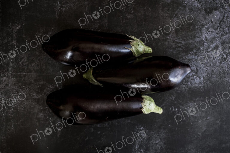 Three glossy purple aubergine egg plant vegetables on a grey slate background