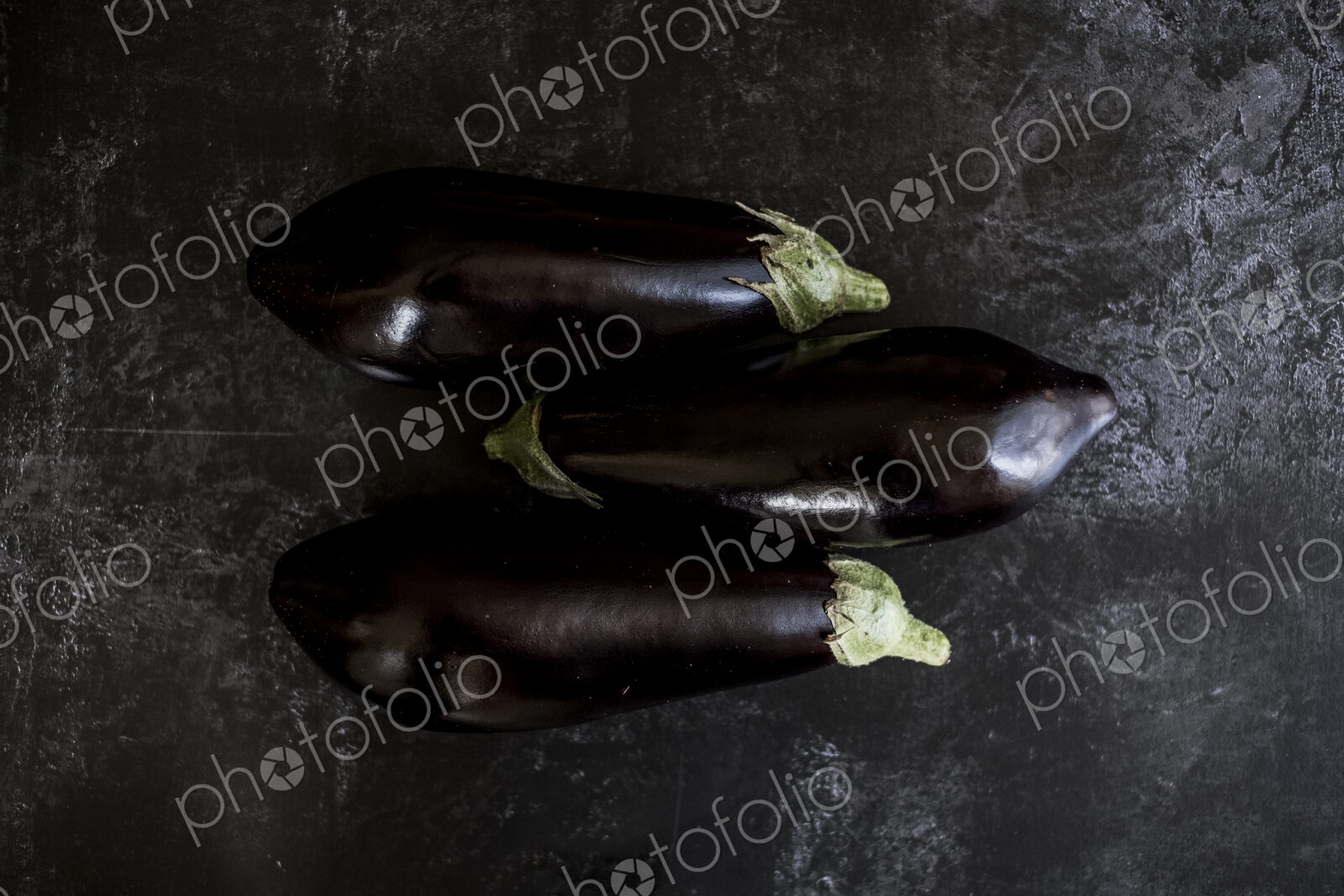 Three glossy purple aubergine egg plant vegetables on a grey slate background