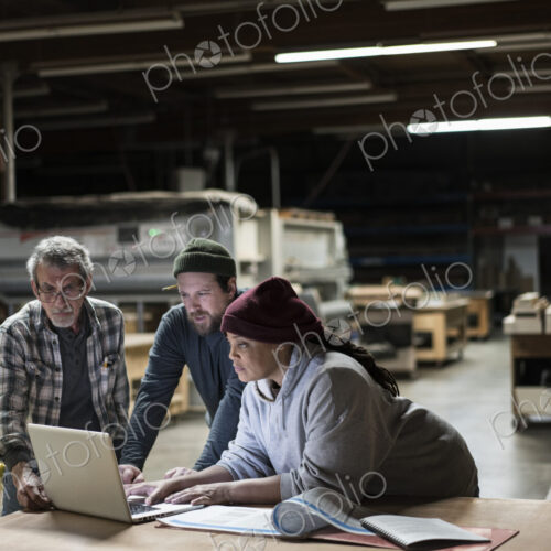Three mixed race carpenters workng on a laptop computer after work hours in a large… Three mixed race carpenters workng on a laptop computer after work hours in a large…