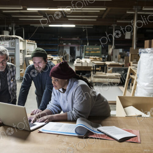 Three mixed race carpenters workng on a laptop computer after work hours in a large…