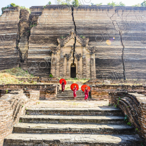 Three monks with orange umbrellas outside a large rock temple, the entrance carved… Three monks with orange umbrellas outside a large rock temple, the entrance carved…
