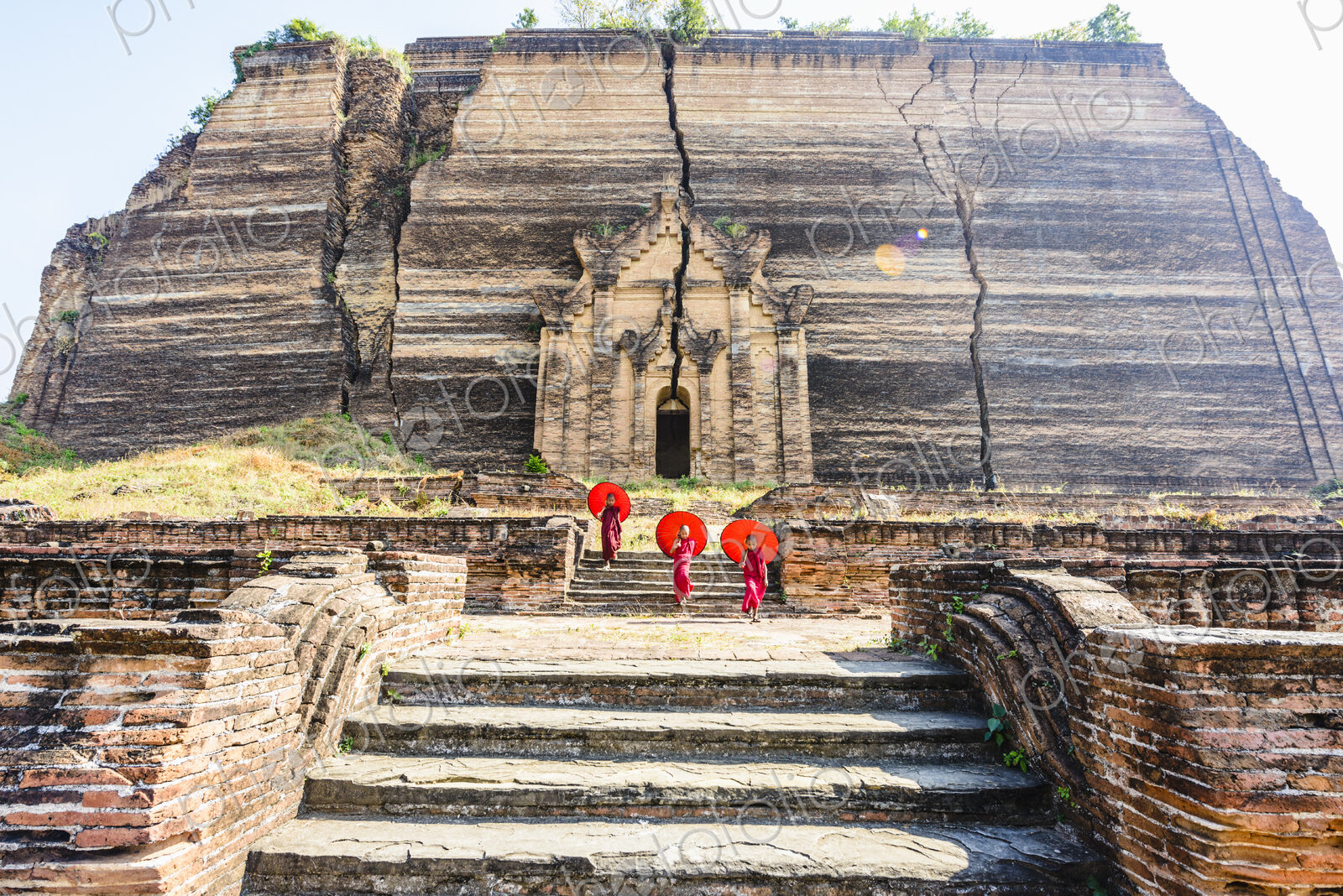 Three monks with orange umbrellas outside a large rock temple, the entrance carved…