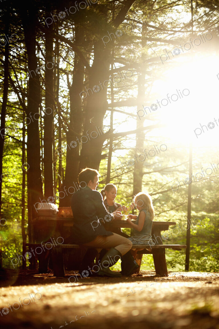 Three people, a family sitting at a picnic table under trees, in the late afternoon.