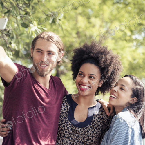 Three people, a man and two women, taking selfies in a city park