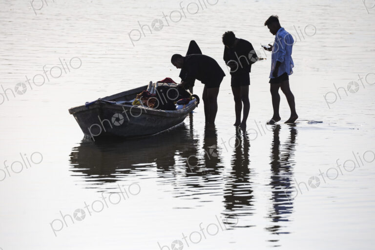 Three people in shallow water, one holding a small wooden boat, silhouettes.