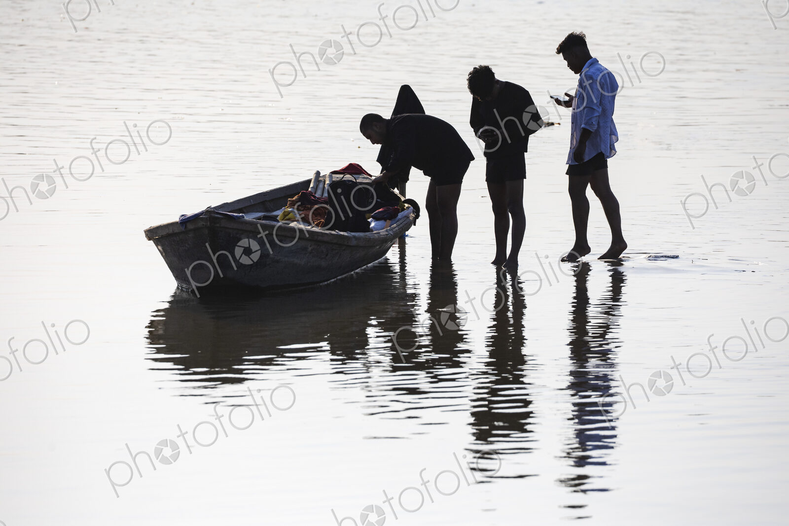 Three people in shallow water, one holding a small wooden boat, silhouettes.
