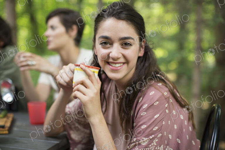 Three people seated around a table in a woodland clearing, one woman smiling at the…
