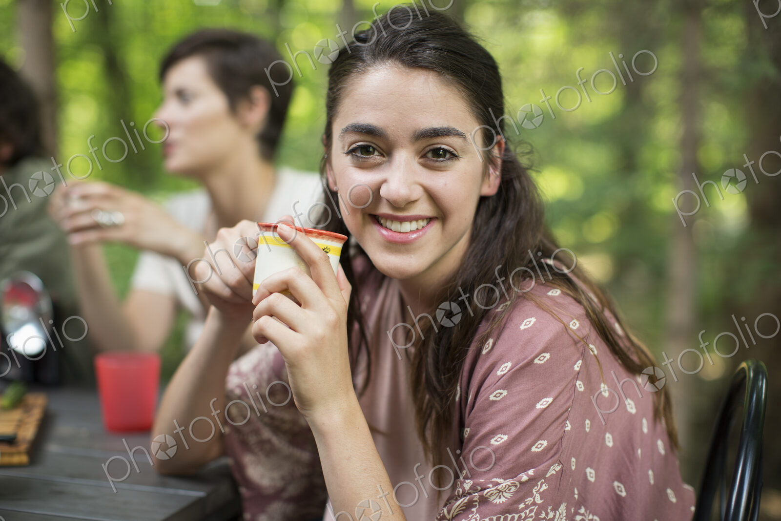 Three people seated around a table in a woodland clearing, one woman smiling at the…