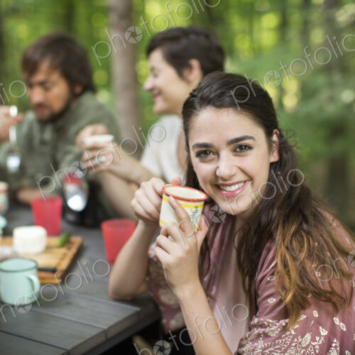 Three people seated around a table in a woodland clearing, one woman smiling at the… Three people seated around a table in a woodland clearing, one woman smiling at the…
