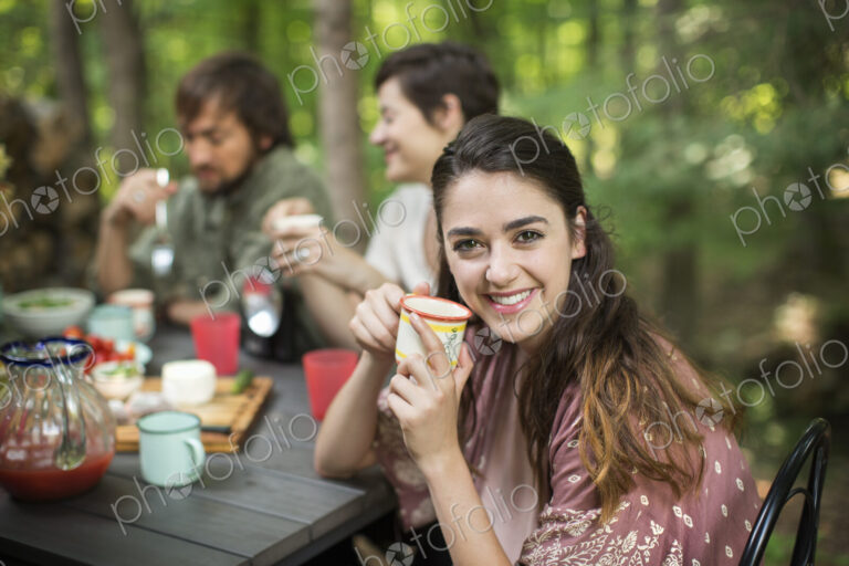 Three people seated around a table in a woodland clearing, one woman smiling at the…