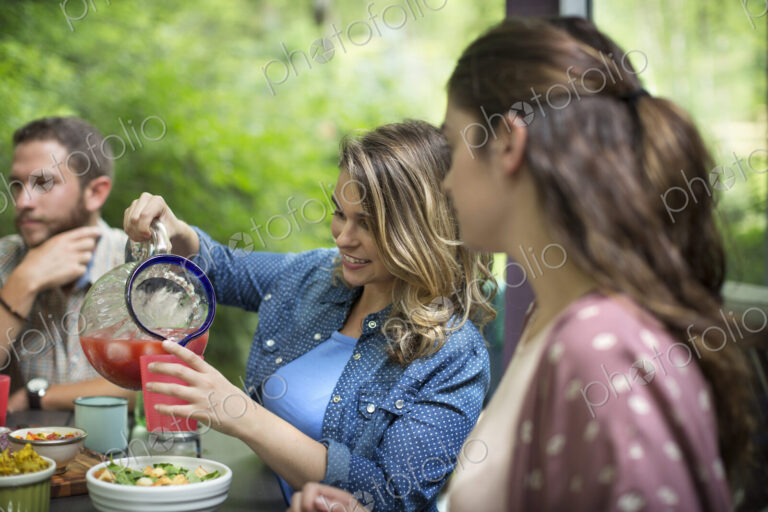 Three people seated at a table in woodland, having a meal, one woman pouring juice…