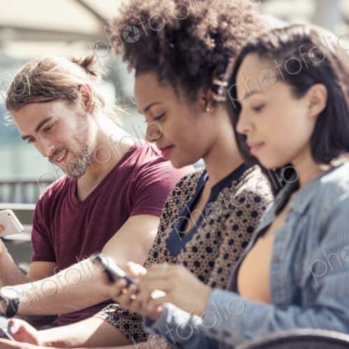 Three people seated side by side on a park bench checking their smart phones Three people seated side by side on a park bench checking their smart phones