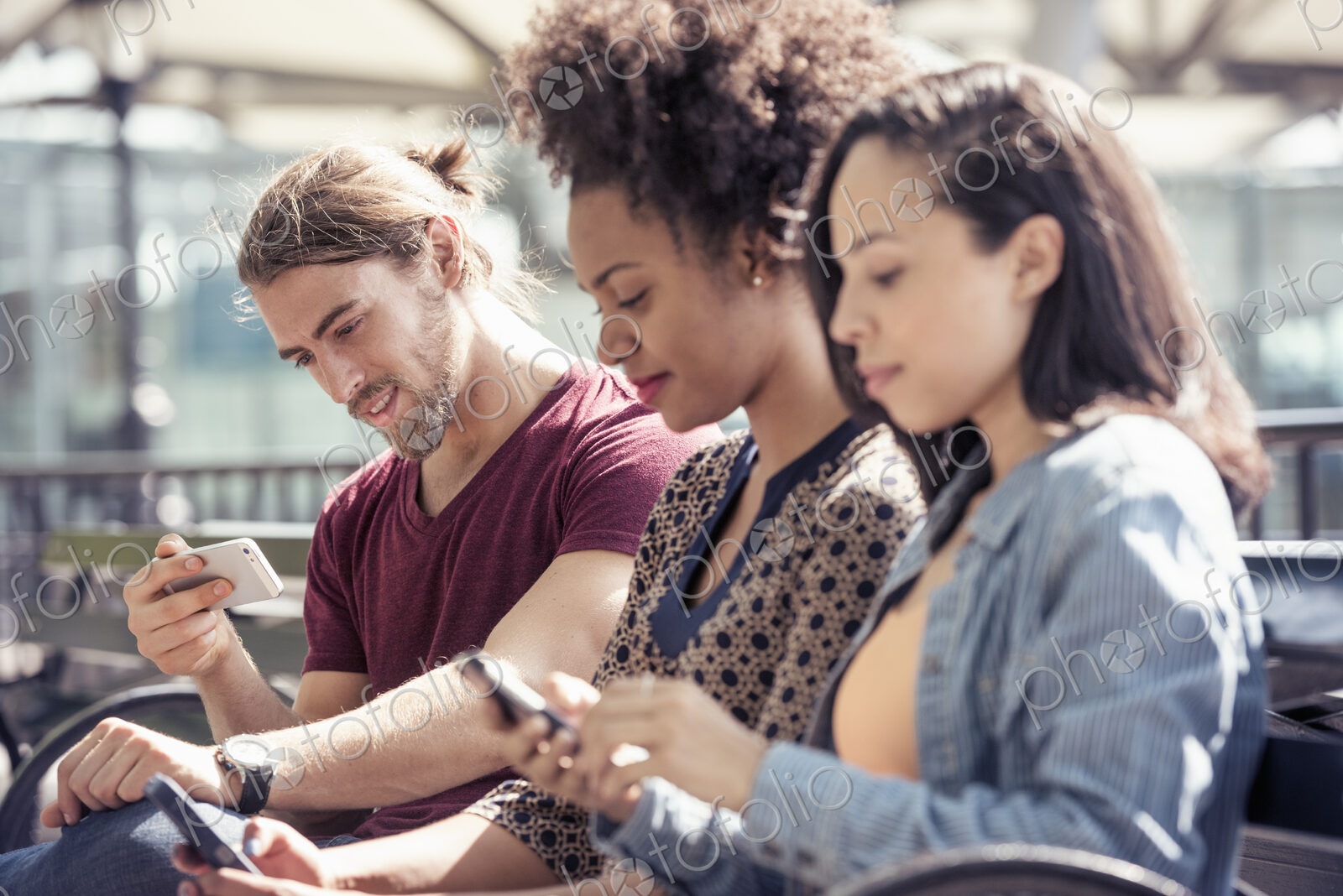 Three people seated side by side on a park bench checking their smart phones