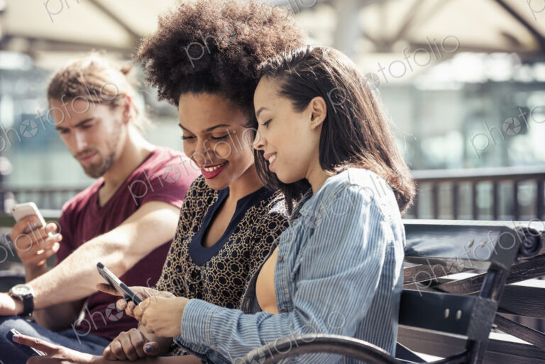 Three people seated side by side on a park bench checking their smart phones
