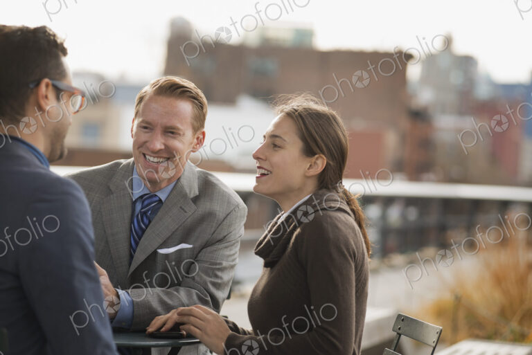 Three people standing in an open space between city buildings, talking to each other.…