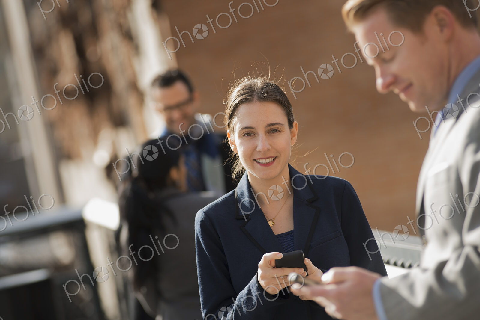 Three people standing on the sidewalk in the city, checking their phones. Two men and…