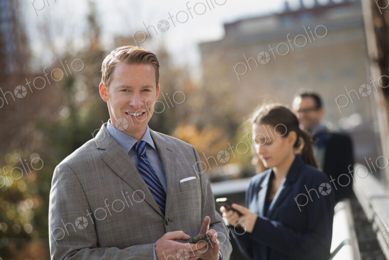 Three people standing on the sidewalk in the city, checking their phones. Two men and…
