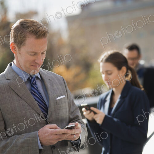 Three people standing on the sidewalk in the city, checking their phones. Two men and… Three people standing on the sidewalk in the city, checking their phones. Two men and…