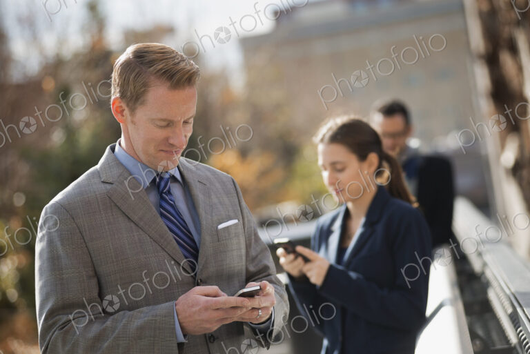 Three people standing on the sidewalk in the city, checking their phones. Two men and…
