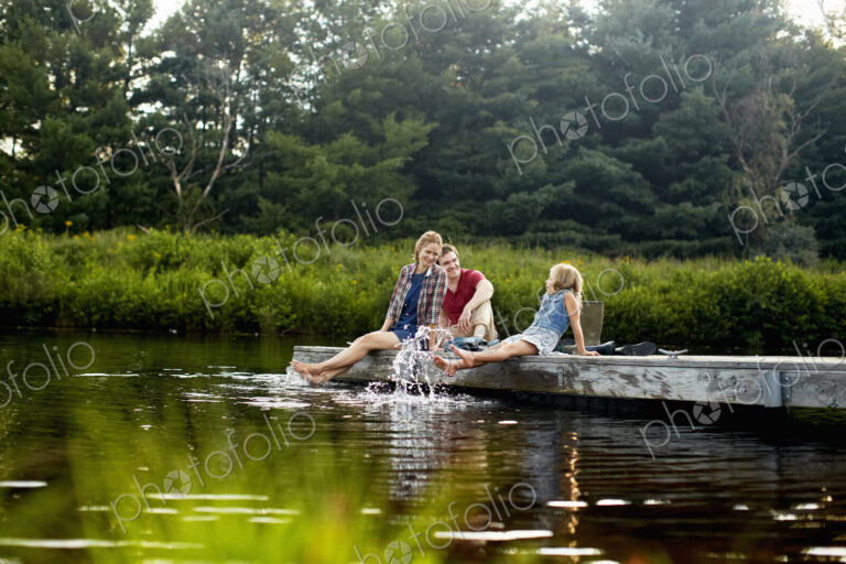 Three people, two adults and a child relaxing on a jetty, with their feet in the water…