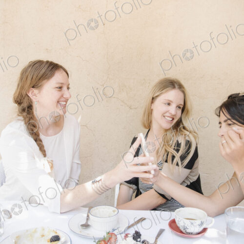 Three smiling young women sitting round a table with food and drink, looking at a cell… Three smiling young women sitting round a table with food and drink, looking at a cell…