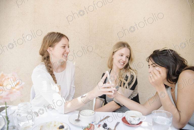 Three smiling young women sitting round a table with food and drink, looking at a cell…