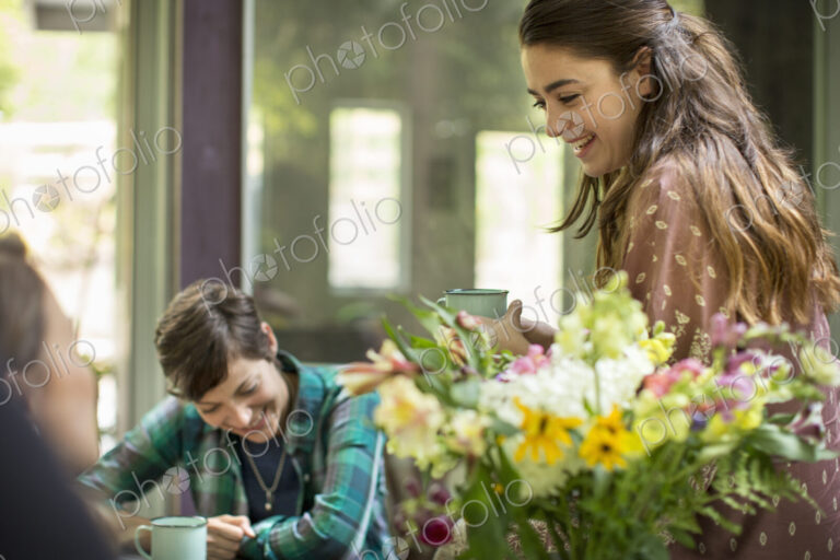 Three women talking and laughing, seated around a table. Large bunch of flowers in a vase.
