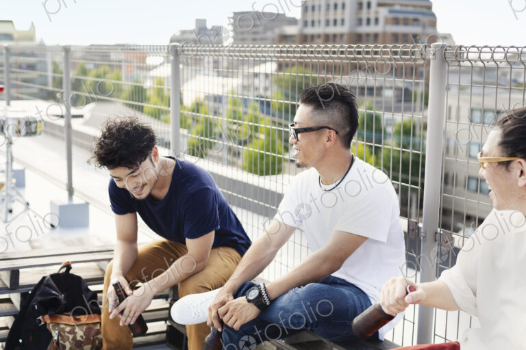 Three young Japanese men sitting on a rooftop in an urban setting, drinking beer.