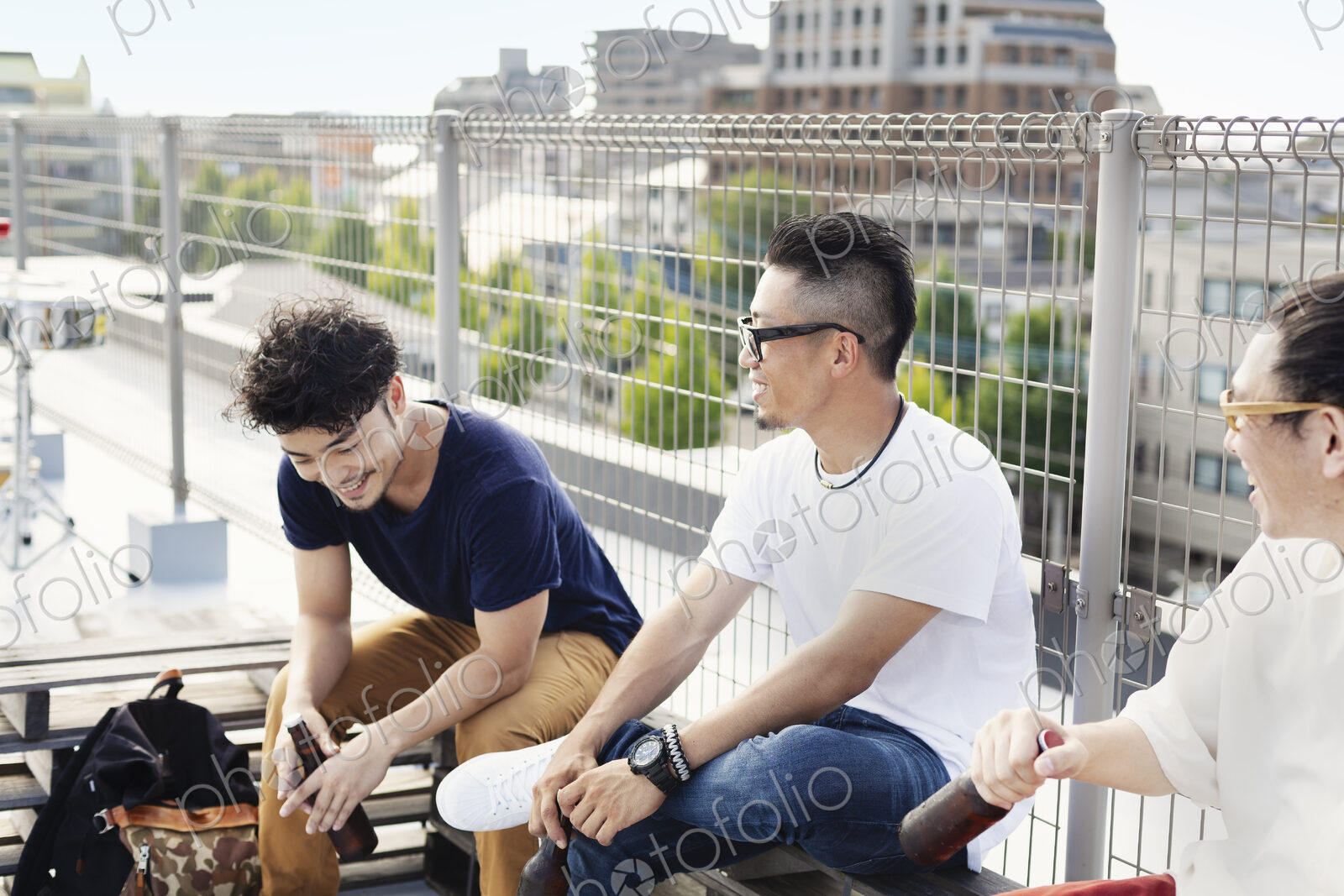 Three young Japanese men sitting on a rooftop in an urban setting, drinking beer.