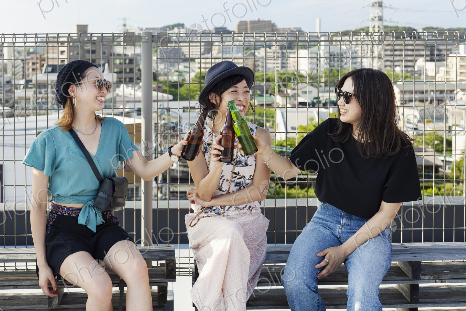 Three young Japanese women sitting on a rooftop in an urban setting, drinking beer.