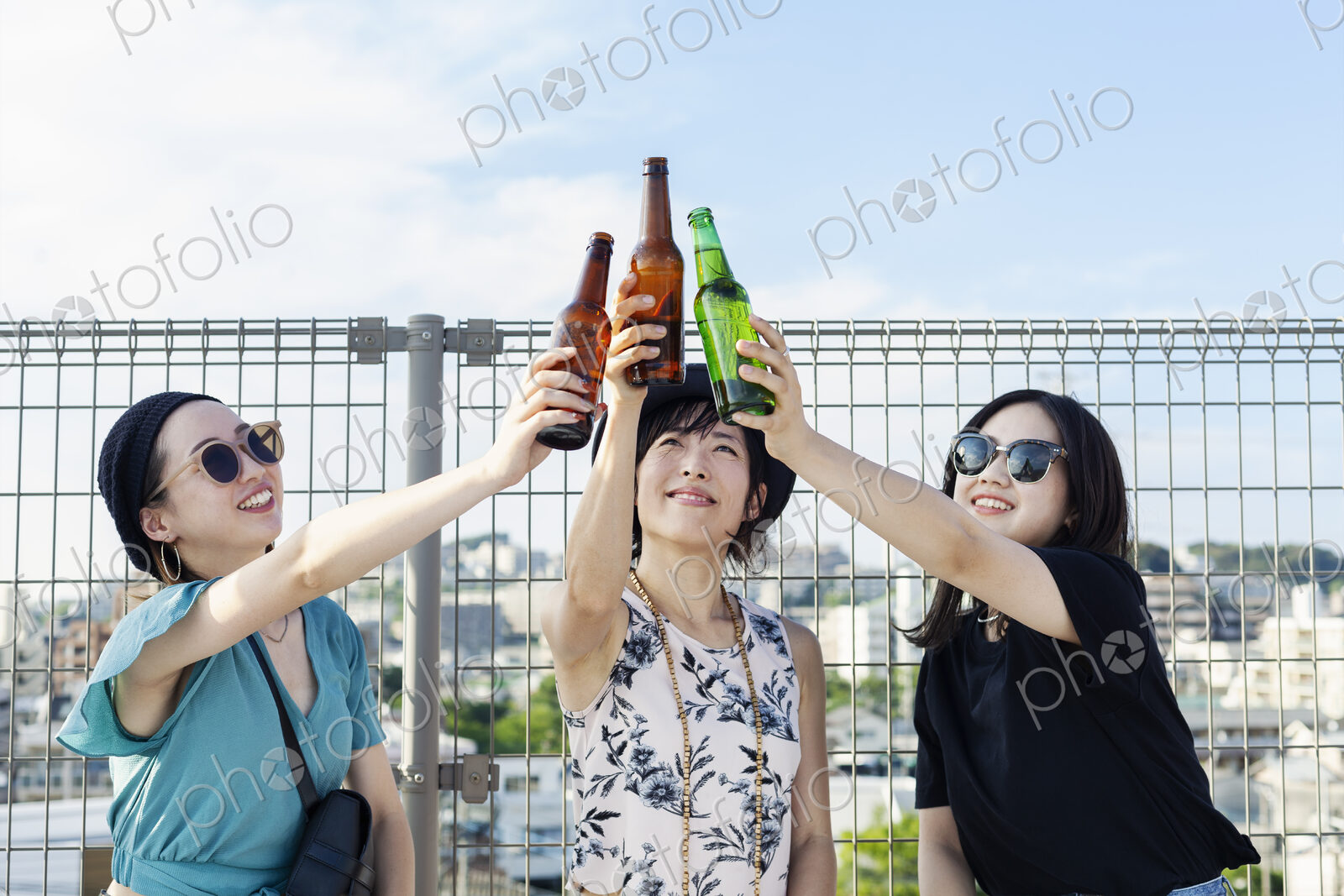 Three young Japanese women sitting on a rooftop in an urban setting, drinking beer.