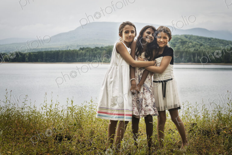 Three young girls standing by the side of a lake, hugging each other.