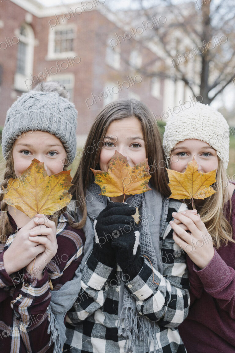 Three young girls standing in a row, holding autumn leaves in front of their faces.