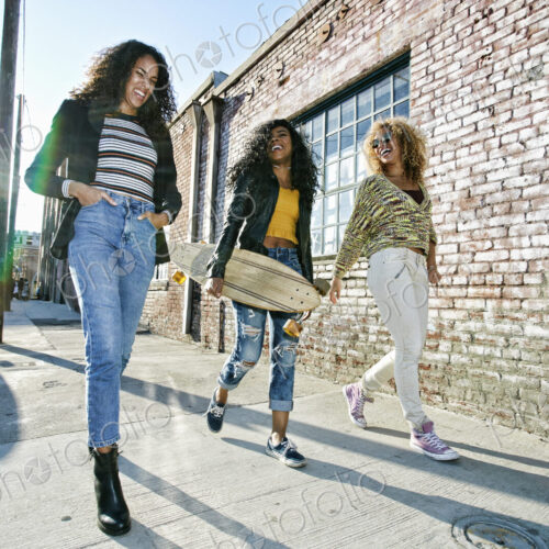 Three young women with long curly hair walking along pavement, one carrying skateboard. Three young women with long curly hair walking along pavement, one carrying skateboard.