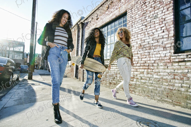 Three young women with long curly hair walking along pavement, one carrying skateboard.