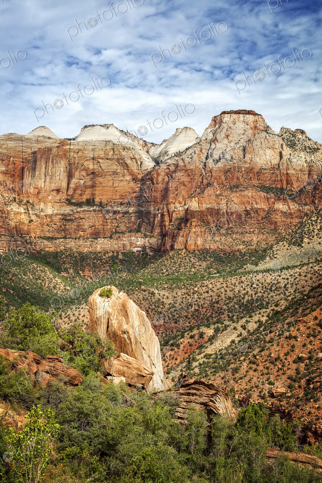 Towering red and white sandstone cliffs rise above rugged desert terrain and green…