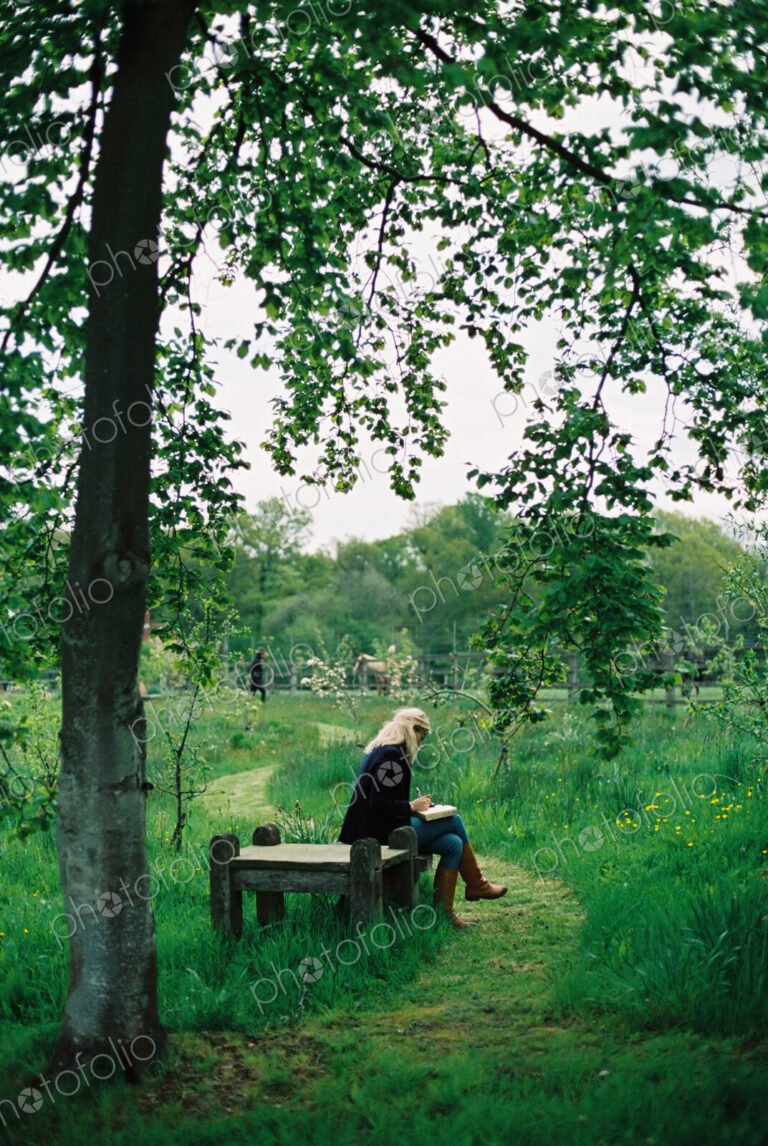 Trees and long grass. A mown path. A woman seated on a wooden seat.