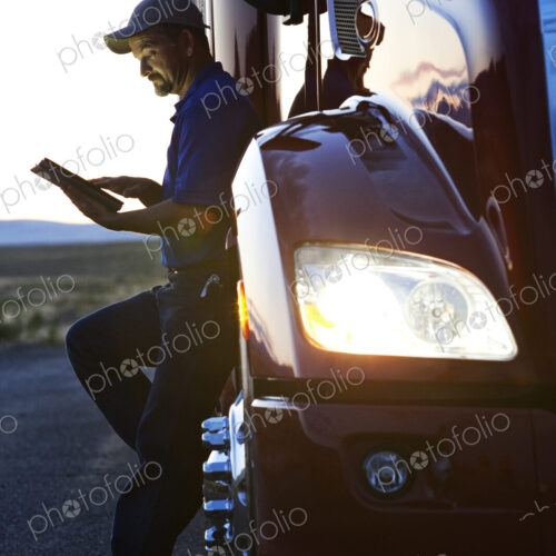 Truck driver checking his notebook computer log while standing next to the cab of his… Truck driver checking his notebook computer log while standing next to the cab of his…