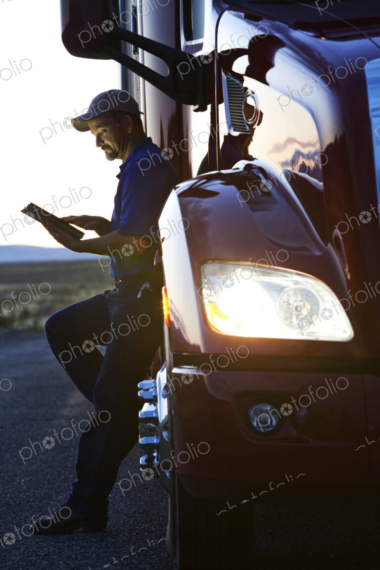 Truck driver checking his notebook computer log while standing next to the cab of his…
