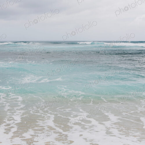 Turquoise ocean water, waves on the beach and an overcast sky.