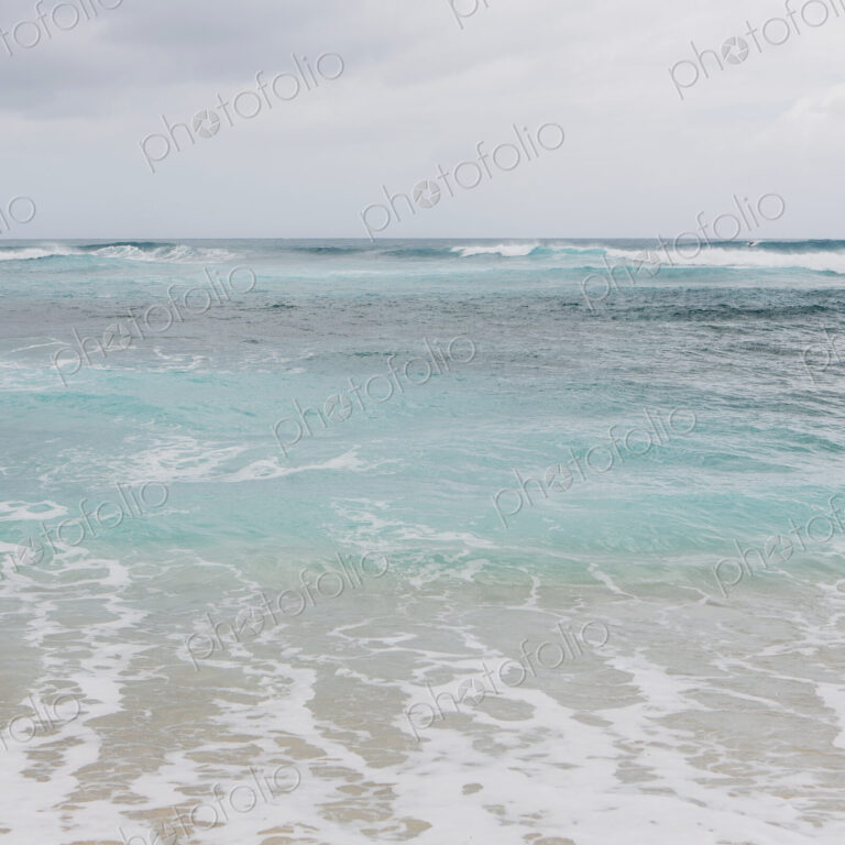 Turquoise ocean water, waves on the beach and an overcast sky.