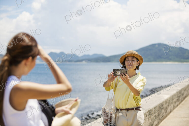 Two Japanese women standing by the ocean, taking picture with mobile phone.