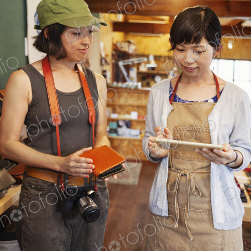 Two Japanese women standing in a leather shop, holding leather purse and digital tablet.