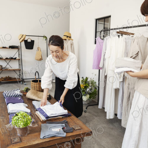 Two Japanese women standing in a small fashion boutique, arranging T-Shirts on a…