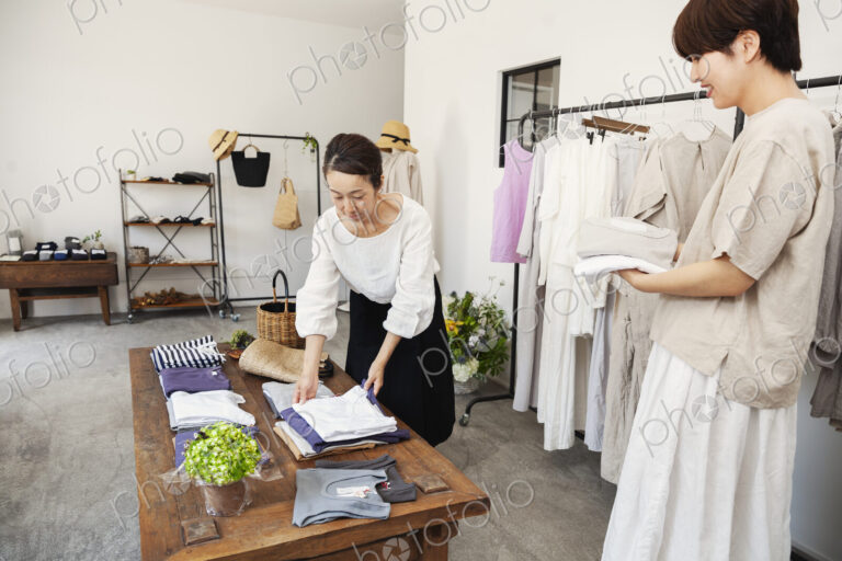 Two Japanese women standing in a small fashion boutique, arranging T-Shirts on a…