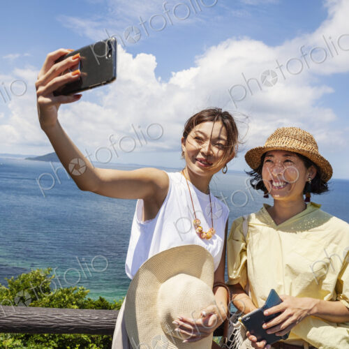 Two Japanese women wearing hats standing on a cliff, taking selfie with mobile phone,… Two Japanese women wearing hats standing on a cliff, taking selfie with mobile phone,…