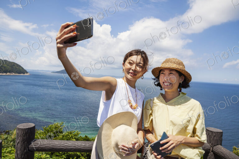 Two Japanese women wearing hats standing on a cliff, taking selfie with mobile phone,…
