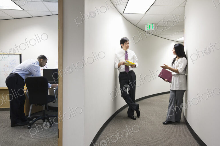 Two asian businesspeople talking in a hallway near a black businessman working alone…