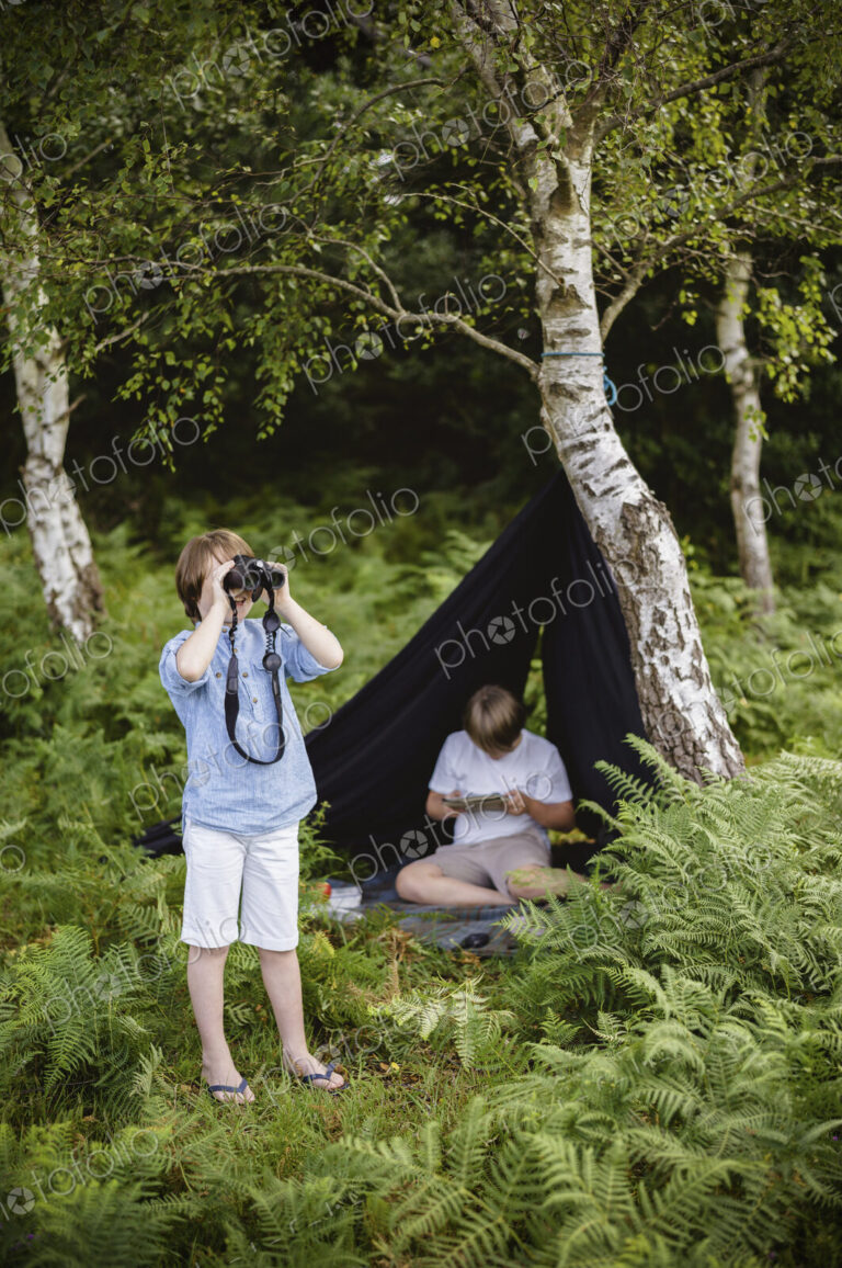 Two boys camping in New Forest. One sitting under a black canvas shelter. One boy…