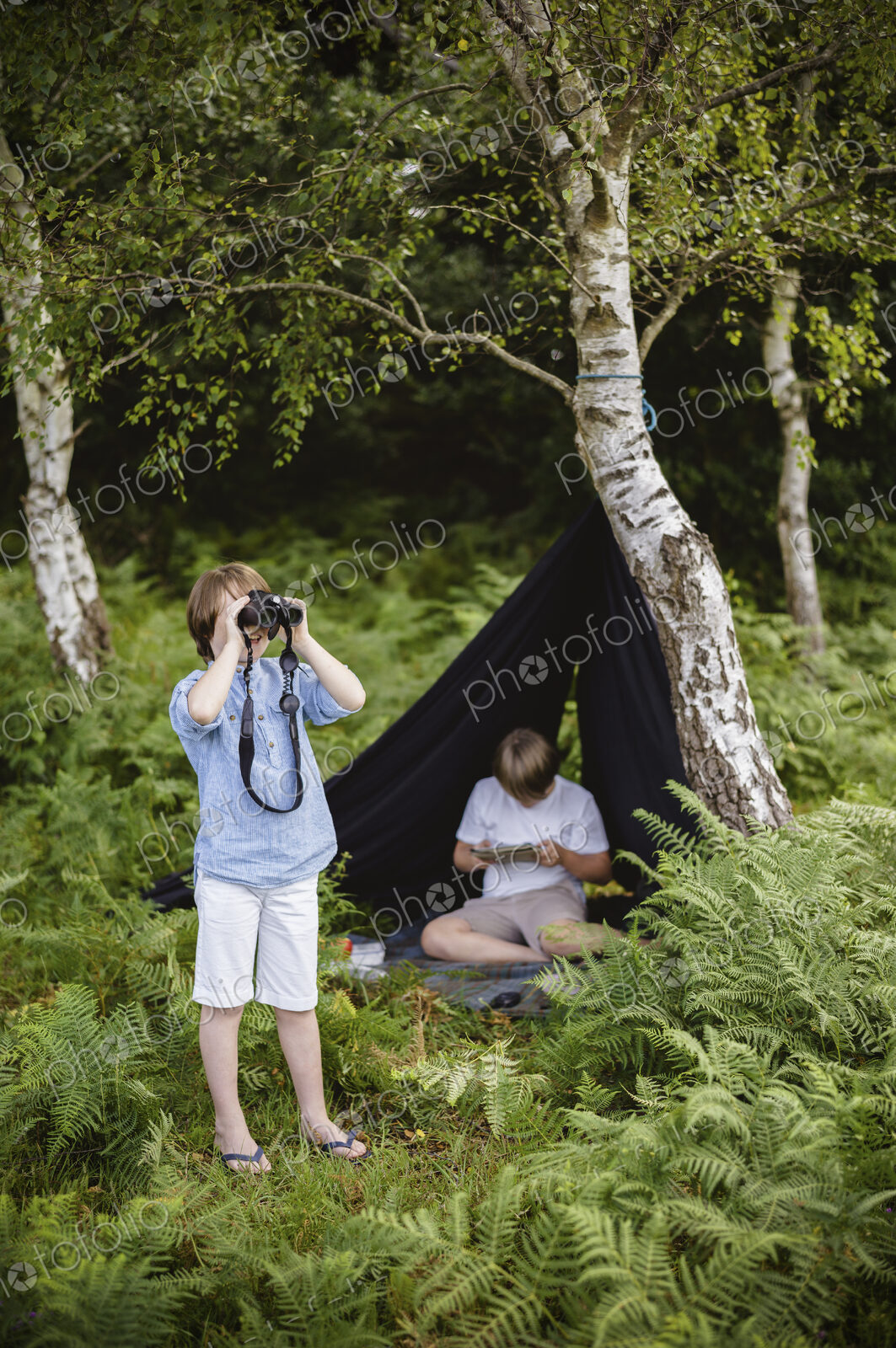 Two boys camping in New Forest. One sitting under a black canvas shelter. One boy…